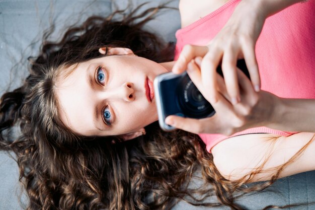 Foto jonge vrouw met lang krullend haar ligt op de bank en scrolt sociale netwerken op haar telefoon