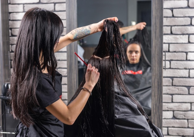 Jonge vrouw met lang nat haar die op kapsel wacht terwijl professionele kapper haar haar kamt, zittend in een fauteuil in schoonheidssalon