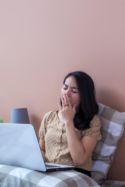 Jonge vrouw met laptop in bed die met handen die mond bedekken geeuwen