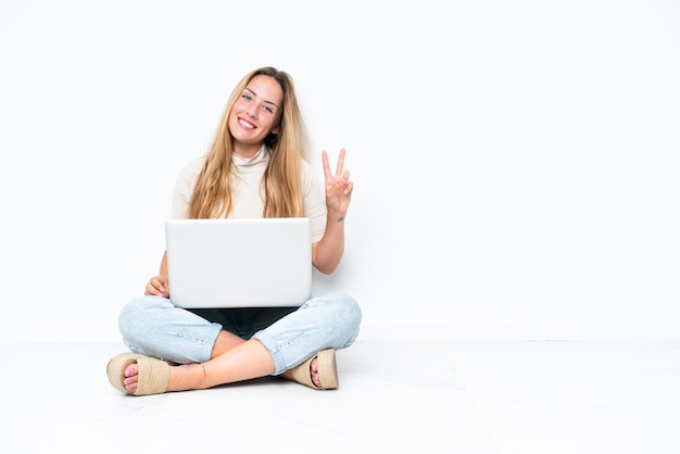Jonge vrouw met laptop zittend op de vloer geïsoleerd op een witte achtergrond glimlachend en overwinning teken tonen