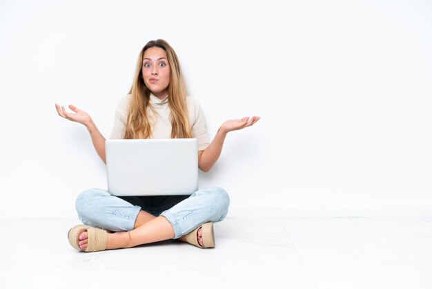 Jonge vrouw met laptop zittend op de vloer geïsoleerd op een witte achtergrond met twijfels tijdens het opsteken van handen