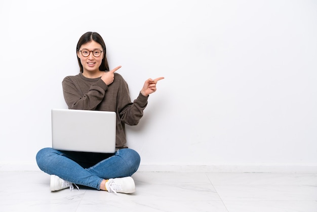 Jonge vrouw met laptop zittend op de vloer wijzende vinger naar de zijkant en presenteert een product