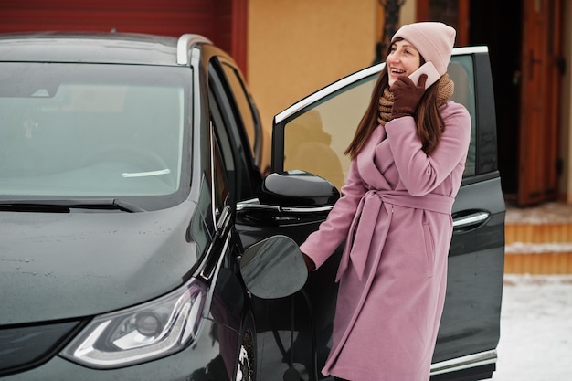 Jonge vrouw met mobiele telefoon bij de hand die haar elektrische auto in de winter oplaadt.