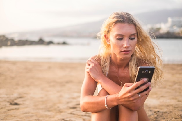 Jonge vrouw met mobiele telefoon terwijl ze op het strand tegen de lucht zit