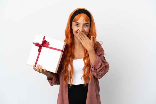 Jonge vrouw met oranje haar die een geschenk vasthoudt dat op een witte achtergrond wordt geïsoleerd, gelukkig en glimlachend die de mond bedekt met de hand