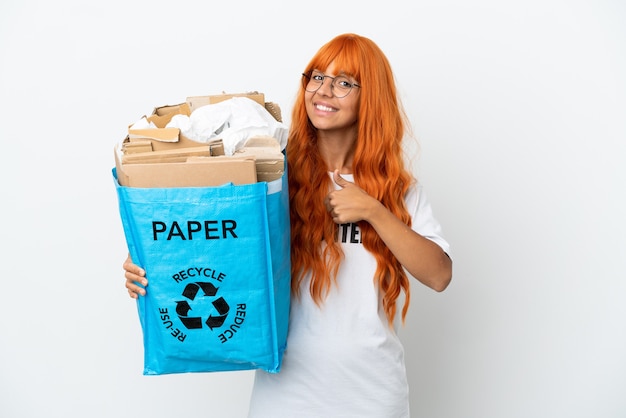 Jonge vrouw met oranje haar met een recyclingzak vol papier om te recyclen geïsoleerd op een witte achtergrond en geeft een duim omhoog gebaar