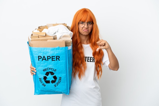 Jonge vrouw met oranje haar met een recyclingzak vol papier om te recyclen geïsoleerd op een witte achtergrond met duim omlaag met negatieve uitdrukking