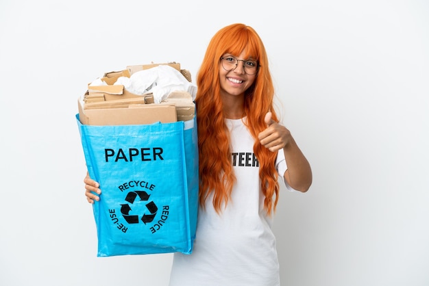 Jonge vrouw met oranje haar met een recyclingzak vol papier om te recyclen geïsoleerd op een witte achtergrond met duimen omhoog omdat er iets goeds is gebeurd