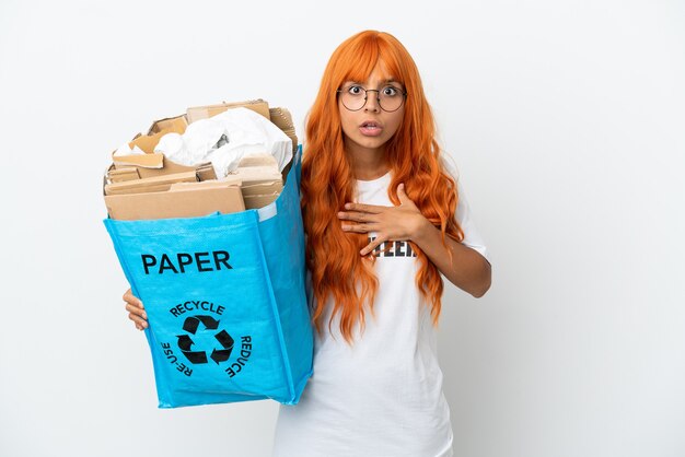 Jonge vrouw met oranje haar met een recyclingzak vol papier om te recyclen geïsoleerd op een witte achtergrond verrast en geschokt terwijl ze er goed uitziet