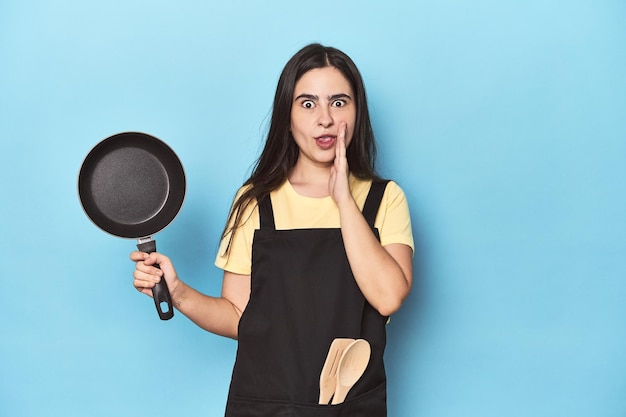 Jonge vrouw met pan op blauwe studio zegt een geheim heet remnieuws en kijkt opzij