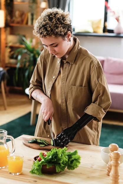 Jonge vrouw met prothetische arm die verse groente aan tafel snijdt en salade kookt voor de lunch