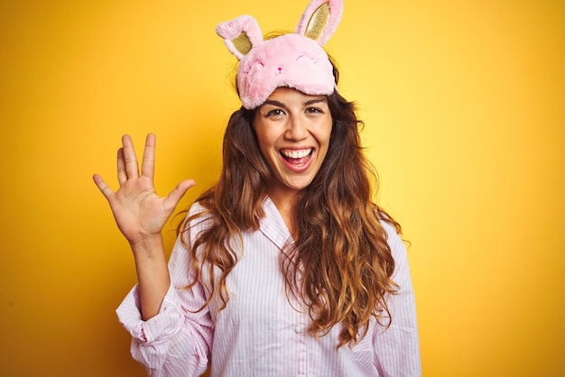 Jonge vrouw met pyjama en slaapmasker die over een gele geïsoleerde achtergrond staat en omhoog wijst met vingers nummer vijf terwijl ze zelfverzekerd en gelukkig glimlacht