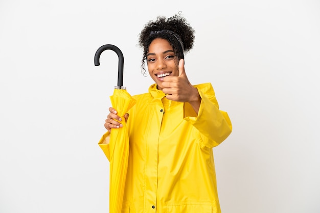 Jonge vrouw met regenbestendige jas en paraplu geïsoleerd op een witte achtergrond met duimen omhoog omdat er iets goeds is gebeurd