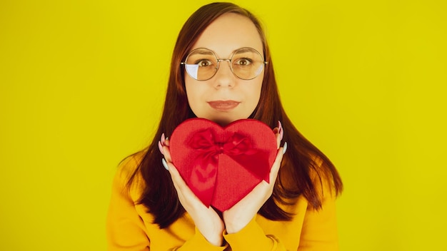 Jonge vrouw met rode geschenkdoos in de vorm van een hart op gele achtergrond Mooie vrouw houdt romantisch cadeau camera kijken en glimlachen Concept van heden op valentijnsdag en internationale vrouwendag