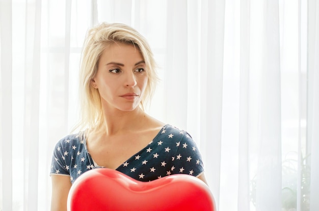 Jonge vrouw met rode hartvormige ballon tegen het raam thuis. Het vieren van Valentijnsdag, bruiloft en liefdesdag, vrouwendag, Wereldhartdag, verjaardag. Afgetekend tegen de zon binnen.