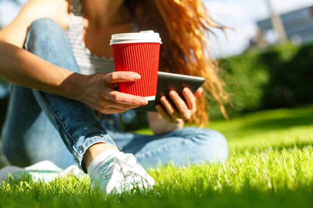 Jonge vrouw met rode wegwerp papieren kopje koffie met behulp van tablet pc op een groen gazon in een park