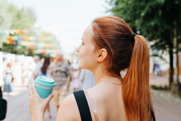 Jonge vrouw met rood haar genieten op straat festival vakantie wegkijken achteraanzicht Mooie vrouw met kopje koffie op outdoor evenement op zomerdag mensen lopen versierde straat in bokeh