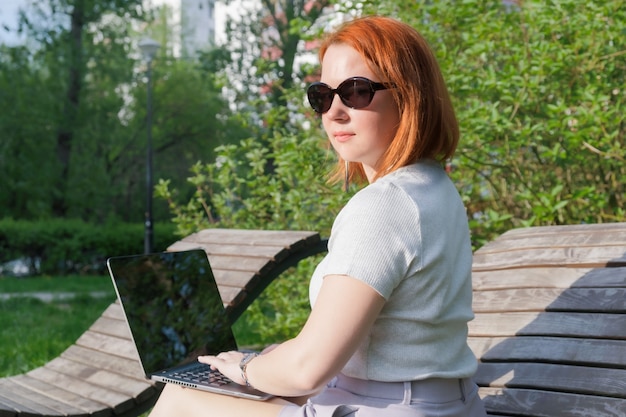 Jonge vrouw met rood haar met een laptop op een bankje. freelancer vrouw werkt in het park. gefotografeerd vanaf de achterkant, draaide de vrouw zich om.