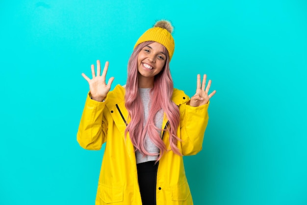 Jonge vrouw met roze haar, gekleed in een regenbestendige jas geïsoleerd op een blauwe achtergrond die negen met vingers telt