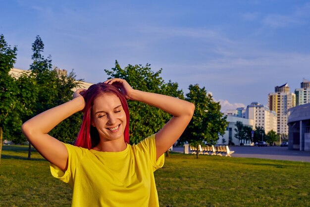 Jonge vrouw met roze haar, gele crop top sloot haar ogen, glimlachend in stadspark