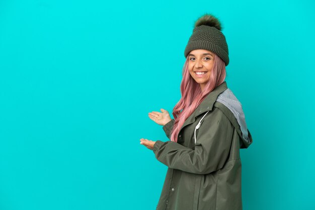 Jonge vrouw met roze haar met een regenbestendige jas geïsoleerd op een blauwe achtergrond die haar handen naar de zijkant uitstrekt om uit te nodigen om te komen
