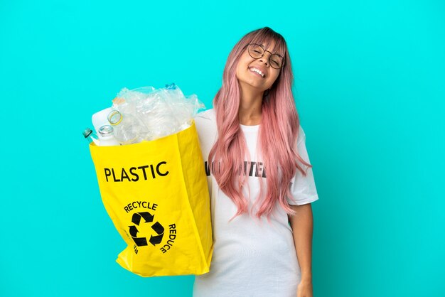 Jonge vrouw met roze haar met een zak vol plastic flessen om te recyclen geïsoleerd op blauwe achtergrond lachen