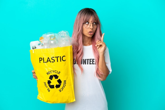 Jonge vrouw met roze haar met een zak vol plastic flessen om te recyclen geïsoleerd op een blauwe achtergrond, denkend aan een idee met de vinger omhoog