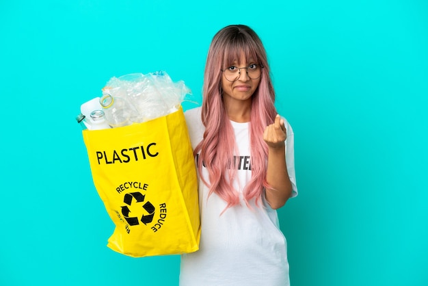 Jonge vrouw met roze haar met een zak vol plastic flessen om te recyclen, geïsoleerd op een blauwe achtergrond, geld gebaar maken