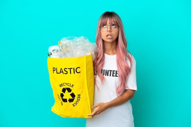 Jonge vrouw met roze haar met een zak vol plastic flessen om te recyclen geïsoleerd op een blauwe achtergrond, omhoog kijkend en met een verbaasde uitdrukking