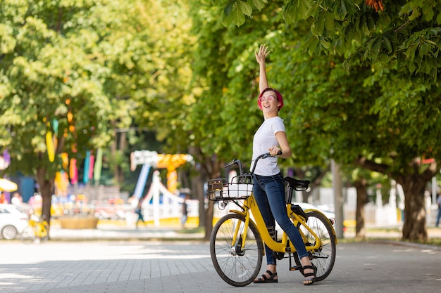 Jonge vrouw met roze haren loopt in het park met een fiets die ijs eet in de zomer.