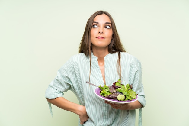 Jonge vrouw met salade over geïsoleerde groene muur die twijfelsgebaar maken terwijl het opheffen van de schouders