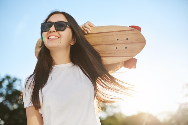 Jonge vrouw met skateboard of longboard die een zonnebril draagt op een zonnige zomerdag in het park