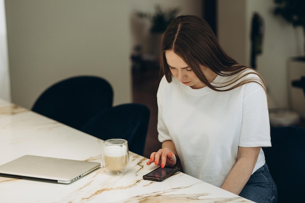 Jonge vrouw met smartphone leunend aan de keukentafel met koffiemok en organisator in een modern huis Glimlachende vrouw die telefoonbericht leest Brunette gelukkig meisje een SMS-bericht te typen