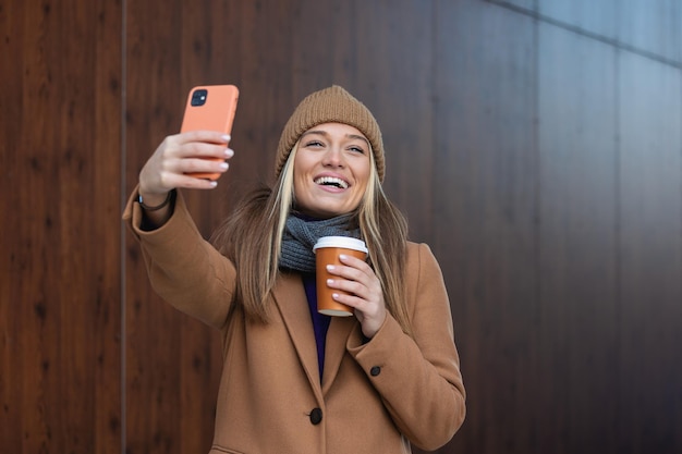 Jonge vrouw met smartphone staande tegen straat wazig gebouw achtergrond Mode foto van mooi meisje met telefoon en kopje koffie