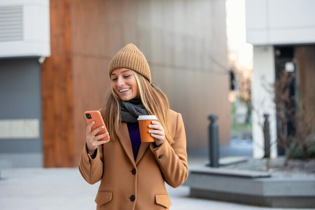 Jonge vrouw met smartphone staande tegen straat wazig gebouw achtergrond Mode zakelijke foto van mooi meisje met telefoon en kopje koffie