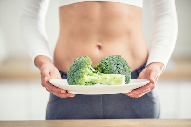 Jonge vrouw met sportfiguur in de keuken met in haar handen en een bord broccoli.