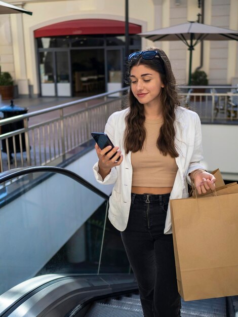 Jonge vrouw met tassen en mobiele telefoon die de roltrap opgaat naar het winkelcentrum