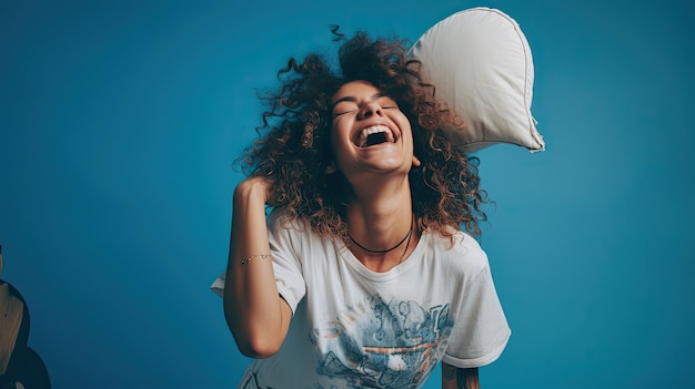 jonge vrouw met tatoeages en krullend haar die een slaapmasker draagt en gelukkig springt terwijl ze een kussen vasthoudt tegen een levendige blauwe achtergrond