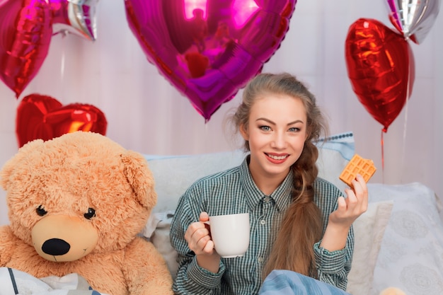 Jonge vrouw met teddybeer ontbijten met wafels en thee of koffie op bed op Valentijnsdag onder ballonnen