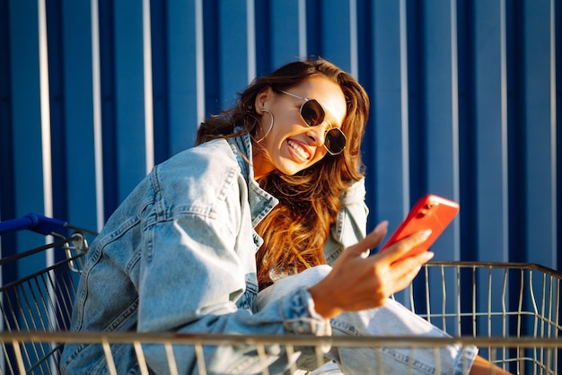 Jonge vrouw met telefoon zitten winkelwagentje plezier in de buurt van kleur muur Shopaholic klant