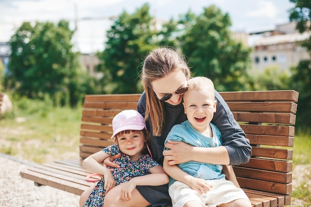 Jonge vrouw met twee kinderen - een jongen en een meisje van 3-4 jaar, zittend op een bankje in het park. Gelukkig jonge moeder met kinderen knuffelen en dwaas spelen. Gelukkige familie tijd samen buiten doorbrengen.