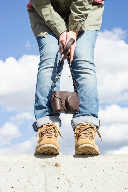 Jonge vrouw met voeten gekleed in laarzen staande op een betonnen muur met een camera in haar handen