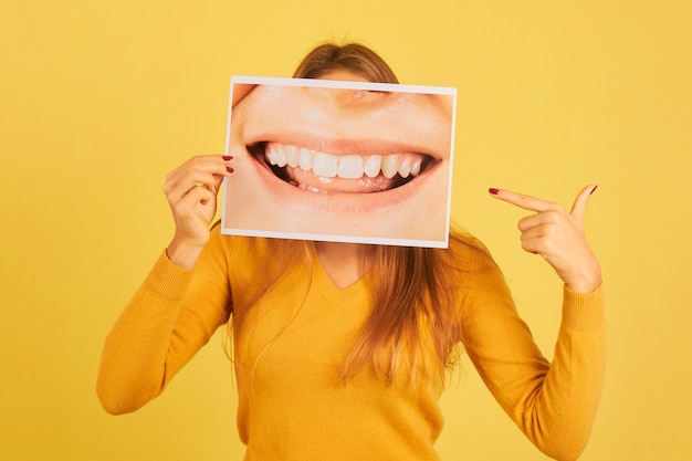 Jonge vrouw met wijzende vinger op foto van mond lachend met haar tanden op gele achtergrond. Tandarts concept