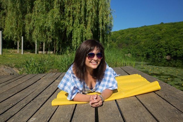 Jonge vrouw met zonnebril ligt op een zonnige dag op een houten strand in de buurt van de rivier en glimlacht