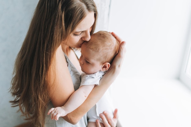 Jonge vrouw moeder met lang haar met babymeisje op handen in de buurt van raam thuis