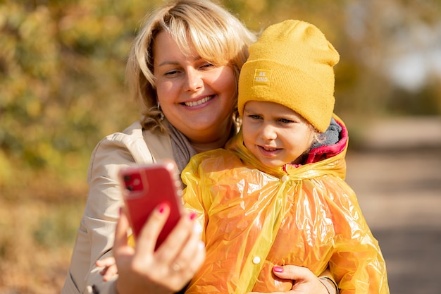 Jonge vrouw, moeder van een klein meisje, gebruikt smartphone om te videobellen met haar familieleden of neemt selfie midden in het herfstpark