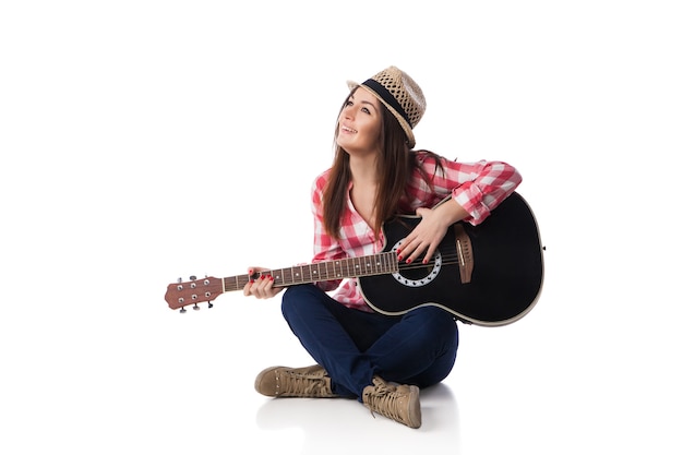 Jonge vrouw muzikant met gitaar dragen shirt en hoed zittend op een vloer en glimlachen. Witte achtergrond.