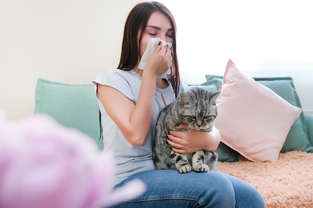 Jonge vrouw niest van bontallergie op de bank en speelt met haar kat.