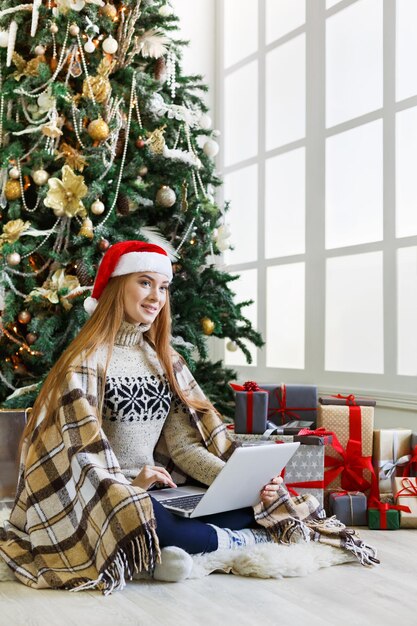 Jonge vrouw online winkelen op laptop in kerst interieur. Meisje zit onder versierde dennenboom tussen veel ingepakte cadeautjes. Voorbereiden op Kerstmis, op winterverkoop, kopieer ruimte