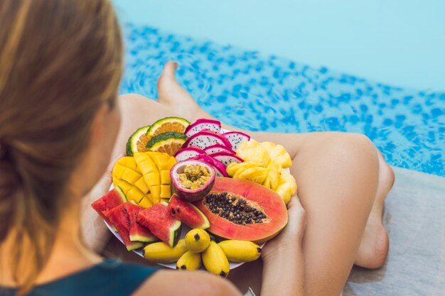 Jonge vrouw ontspannen en eten fruitschaal bij het zwembad van het hotel. Exotisch zomerdieet. Foto van benen met gezond voedsel bij het zwembad, bovenaanzicht van bovenaf. Tropisch strand levensstijl.